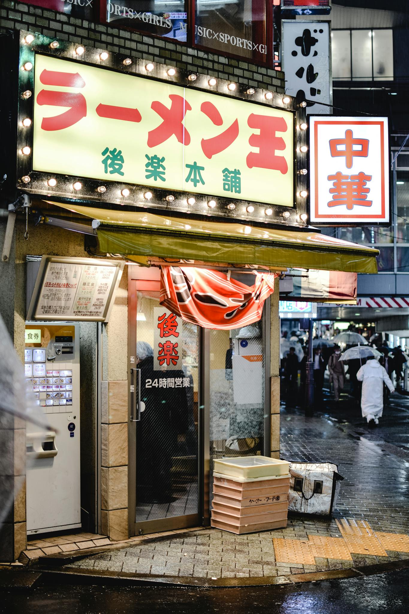 A lively Tokyo street scene with people passing by a ramen shop glowing on a rainy night.