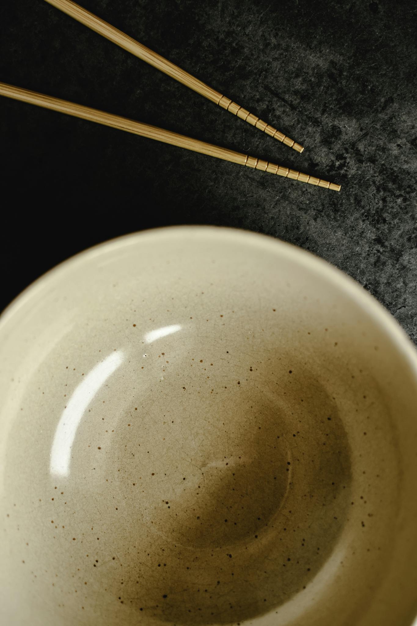 Top view of an empty ceramic bowl and chopsticks on a textured dark surface, creating a minimalist aesthetic.